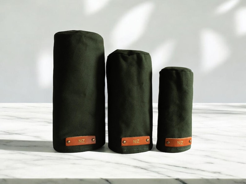 May include: Three dark olive green cylindrical golf club head covers of varying heights. Each cover features a rectangular tan leather patch at the base, secured with two brass rivets, and embossed with the letters NZ in a gold-colored font.