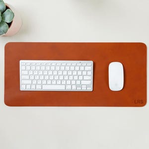 May include: A rectangular, brown leather desk mat with a white keyboard and mouse. A small succulent plant in a pink pot is in the upper left corner. The mat has the initials "LRS" in the lower right corner.