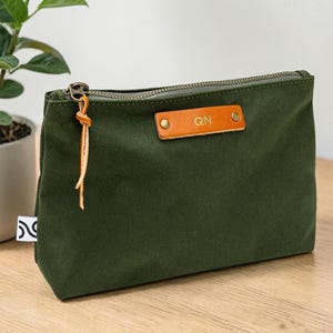 May include: An olive green canvas pouch with a brass zipper and a leather tag. The leather tag has the initials "QN" embossed on it. The pouch is rectangular and sits on a wooden surface. A small plant in a white pot is in the background.