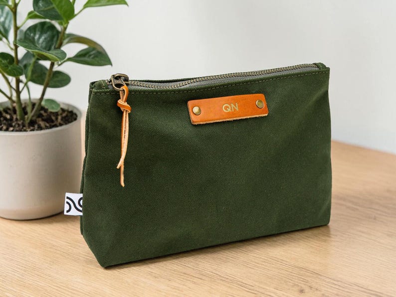 May include: An olive green canvas pouch with a brass zipper and a leather tag. The leather tag has the initials "QN" embossed on it. The pouch is rectangular and sits on a wooden surface. A small plant in a white pot is in the background.