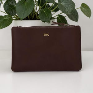 Pode incluir: Uma bolsa de couro castanho escuro com fecho de correr. As letras douradas "ERA" est&atilde;o na frente. A bolsa &eacute; retangular e est&aacute; sobre uma superf&iacute;cie branca, com uma planta em vaso ao fundo. Mede cerca de 25 cm de largura.
