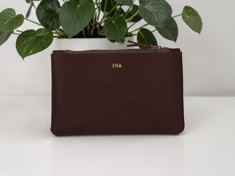 Pode incluir: Uma bolsa de couro castanho escuro com fecho de correr. As letras douradas "ERA" est&atilde;o na frente. A bolsa &eacute; retangular e est&aacute; sobre uma superf&iacute;cie branca, com uma planta em vaso ao fundo. Mede cerca de 25 cm de largura.