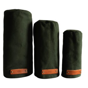 May include: Three cylindrical olive green fabric covers of varying heights, each featuring a rectangular tan leather patch at the base secured with two brass-toned metal rivets. Each leather patch is embossed with a circular logo containing the letters NN.