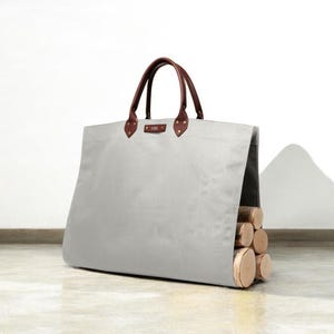 May include: A light grey canvas tote bag with brown leather handles and accents. The bag is filled with several cut logs. The bag has a rectangular shape and is designed for carrying firewood. The bag is sitting on a light-coloured surface.