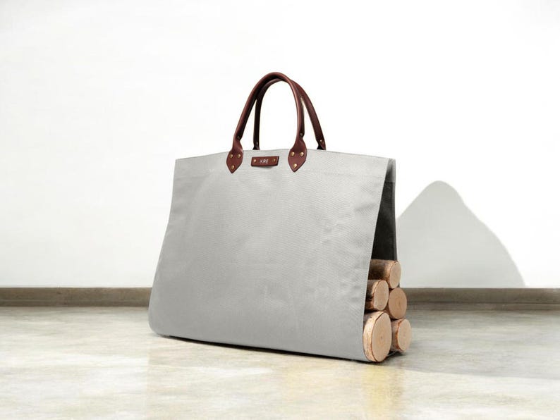 May include: A light grey canvas tote bag with brown leather handles and accents. The bag is filled with several cut logs. The bag has a rectangular shape and is designed for carrying firewood. The bag is sitting on a light-coloured surface.