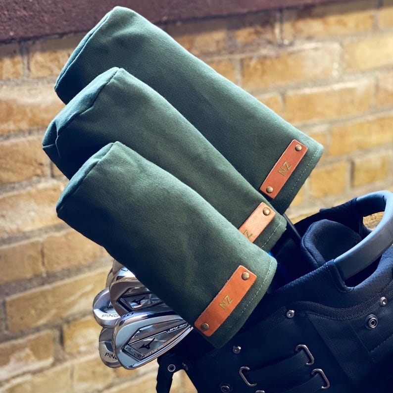 May include: Three olive green canvas golf club head covers with tan leather tabs. Each tab is secured by two brass rivets and features the embossed letters NZ. The covers are fitted over metal golf clubs inside a black golf bag.