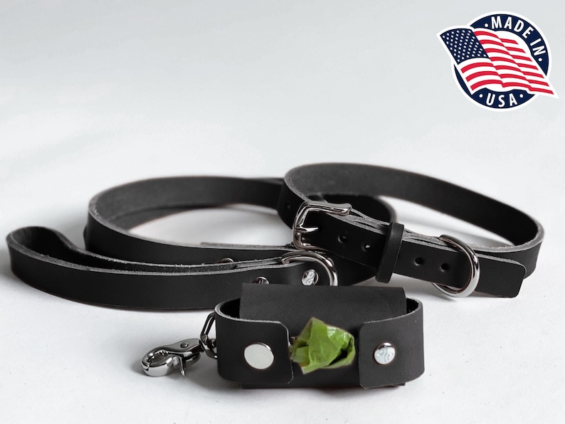 May include: A black leather dog collar and matching leash set with silver-toned metal hardware, accompanied by a small black leather waste bag dispenser featuring a silver snap closure and a green plastic waste bag visible inside.