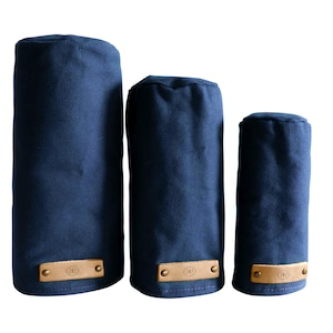 May include: Three navy blue cylindrical golf club head covers of varying heights. Each cover is made of a solid fabric and features a rectangular tan leather patch at the base, secured with two brass-colored metal rivets and a circular logo.