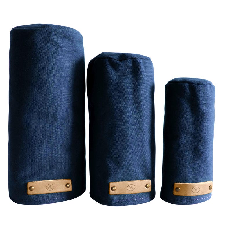 May include: Three navy blue cylindrical golf club head covers of varying heights. Each cover is made of a solid fabric and features a rectangular tan leather patch at the base, secured with two brass-colored metal rivets and a circular logo.