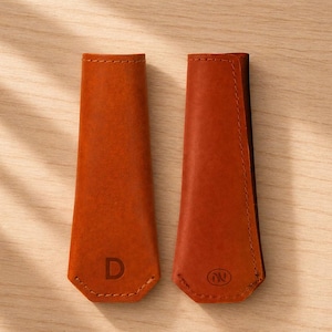 Peut inclure: Deux étuis en cuir brun orangé, probablement pour un couteau ou un outil, sont présentés sur une surface en bois clair. L'un des étuis porte la lettre "D" en relief, l'autre un logo circulaire. Les deux sont cousus.
