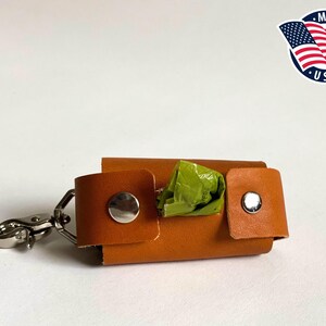 May include: A tan leather waste bag dispenser featuring two silver-toned metal snap buttons and a silver metal swivel lobster clasp. A small portion of a green plastic waste bag is visible protruding from the center of the rectangular holder.