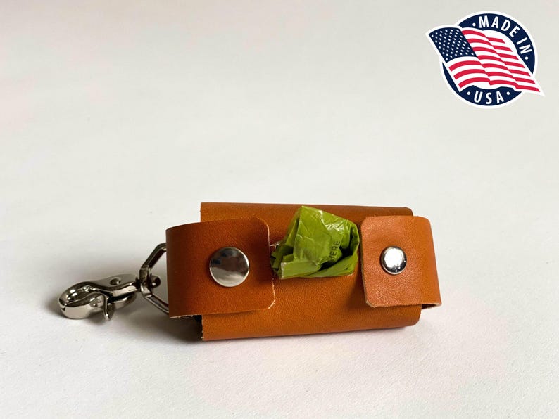 May include: A tan leather waste bag dispenser featuring two silver-toned metal snap buttons and a silver metal swivel lobster clasp. A small portion of a green plastic waste bag is visible protruding from the center of the rectangular holder.