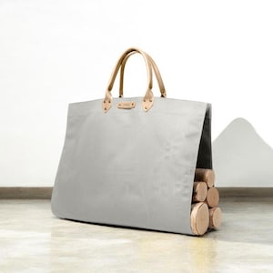 May include: A grey canvas tote bag with light brown leather handles and accents. The bag is filled with several cut logs, showcasing a functional and stylish design. The bag is designed to carry firewood.