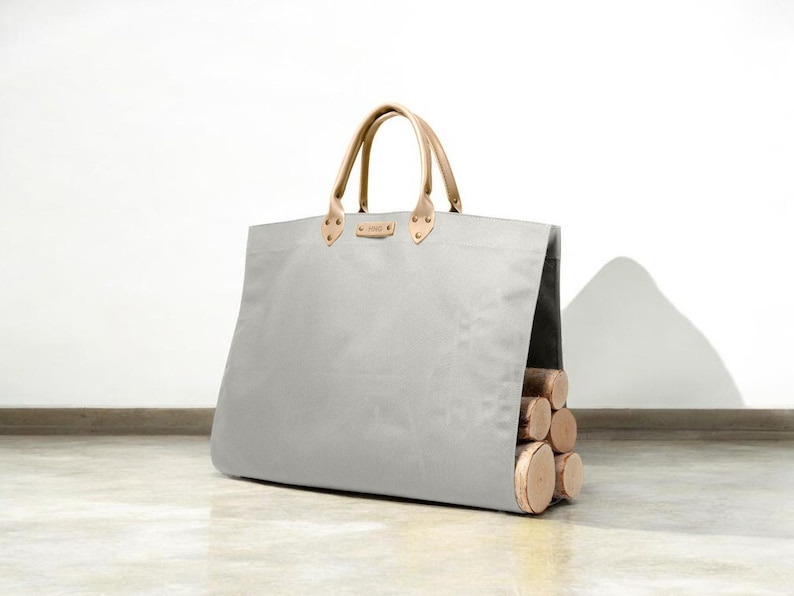 May include: A grey canvas tote bag with light brown leather handles and accents. The bag is filled with several cut logs, showcasing a functional and stylish design. The bag is designed to carry firewood.