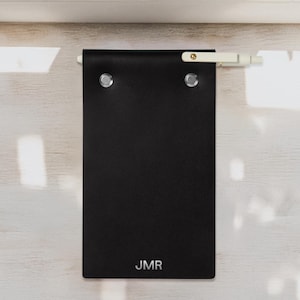 Könnte beinhalten: Ein schwarzes Leder-Notizbuch mit silbernen Nieten und den Initialen "JMR" unten. Das Notizbuch ist an einer weißen Stange mit einer goldenen Schraube befestigt. Der Hintergrund ist eine helle Holzoberfläche.