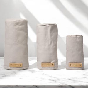 May include: Three cylindrical golf club head covers in a light beige fabric. Each cover features a rectangular tan leather patch at the base, secured with two metal rivets, and embossed with the letters BW in a simple sans-serif font.