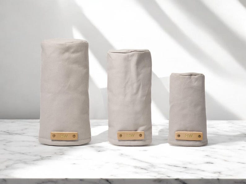 May include: Three cylindrical golf club head covers in a light beige fabric. Each cover features a rectangular tan leather patch at the base, secured with two metal rivets, and embossed with the letters BW in a simple sans-serif font.