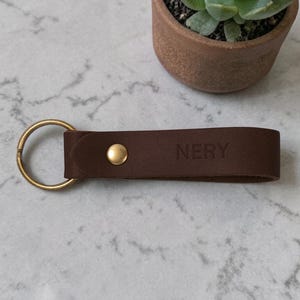 May include: A brown leather keychain with a gold-colored key ring and a gold-colored snap. The word "NERY" is embossed on the leather. A small potted succulent is in the background.