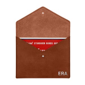 May include: A brown suede envelope-style pouch with a metal snap closure and the letters ERA embossed in the bottom right corner. Inside, a red card is visible with white text reading OFFICIAL STANDARD HANDS AND.