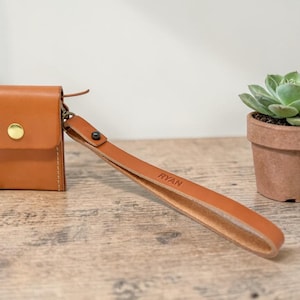 Puede incluir: Una pequeña cartera de cuero marrón con una correa de muñeca. La cartera tiene un cierre de botón dorado y está junto a una pequeña suculenta en maceta. La correa de muñeca está grabada con el nombre "RYAN".