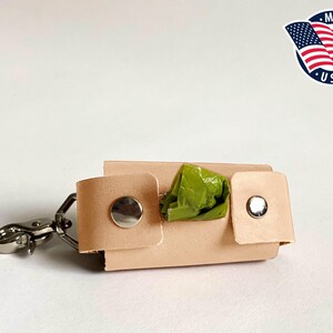 May include: A light tan leather waste bag dispenser featuring two silver-colored metal snap buttons on the front flaps and a silver-colored metal swivel lobster clasp attached to one side. A green plastic waste bag is visible inside the holder.