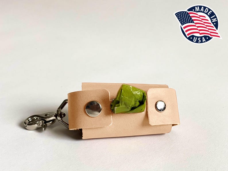 May include: A light tan leather waste bag dispenser featuring two silver-colored metal snap buttons on the front flaps and a silver-colored metal swivel lobster clasp attached to one side. A green plastic waste bag is visible inside the holder.