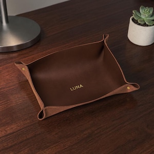 May include: A brown leather catchall tray with the name "LUNA" embossed in gold lettering. The tray has folded corners secured with gold rivets. A small succulent in a white pot sits nearby on a dark wooden surface.