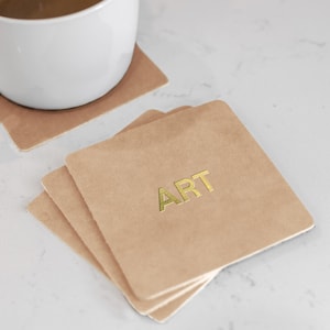 May include: A stack of square, light brown coasters with the word "ART" in gold lettering. A white mug sits on a matching coaster in the background. The coasters are made of a textured material.