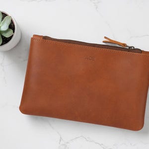 Pode incluir: Uma bolsa de couro castanho com fecho de correr e as letras "ACE" em relevo na frente. Uma pequena planta suculenta num vaso branco est&aacute; no canto superior esquerdo. A bolsa &eacute; retangular.