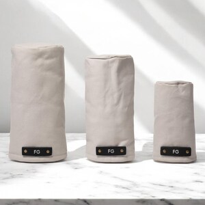 May include: Three cylindrical covers in varying heights made of light beige fabric. Each cover features a rectangular black leather-like tab at the base, secured with two metallic rivets, and embossed with the white capital letters FG.