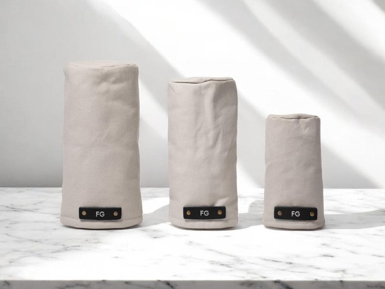 May include: Three cylindrical covers in varying heights made of light beige fabric. Each cover features a rectangular black leather-like tab at the base, secured with two metallic rivets, and embossed with the white capital letters FG.