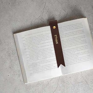 Op de afbeelding: Een bruin leren bladwijzer met de naam "ETHAN" in gouden letters steekt in een open boek. De bladwijzer heeft een gouden klinknagel en een puntige onderkant. Op een grijze ondergrond.