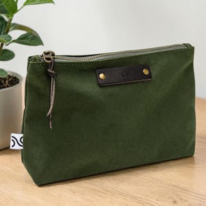 May include: An olive green canvas pouch with a brown leather accent featuring the initials "LV". The pouch has a zipper closure and a leather pull. A small white tag with a black design is visible on the side.