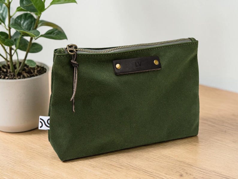 May include: An olive green canvas pouch with a brown leather accent featuring the initials "LV". The pouch has a zipper closure and a leather pull. A small white tag with a black design is visible on the side.