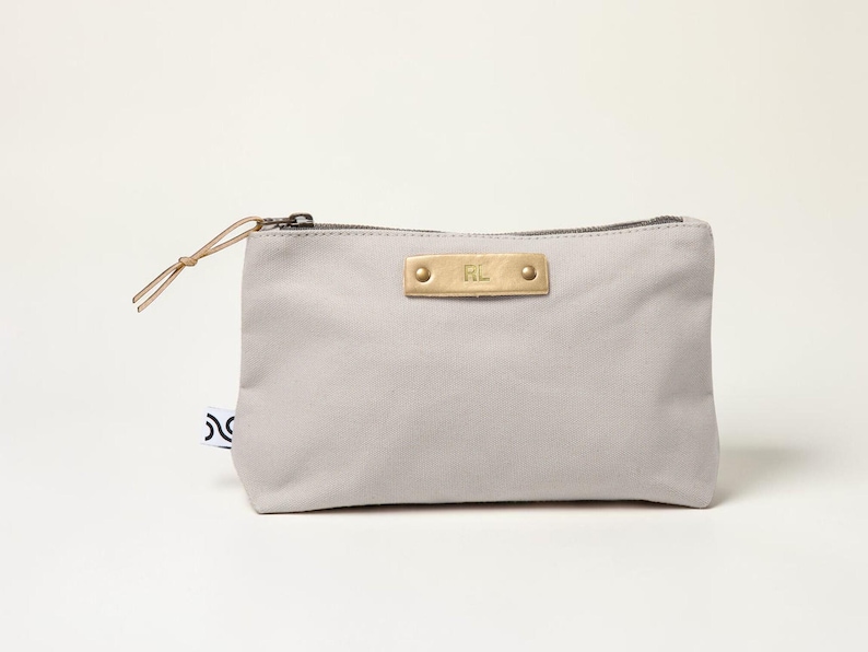 May include: A light gray canvas pouch with a brass nameplate that reads "RL". The pouch has a zipper closure and a leather pull. A small tag with a black and white design is attached to the side.