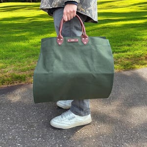 May include: An olive green tote bag with brown leather handles and detailing. The bag is held by a person wearing a camouflage jacket, grey trousers, and white trainers. The bag appears to be made of a durable canvas material.