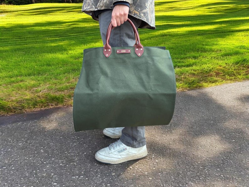May include: An olive green tote bag with brown leather handles and detailing. The bag is held by a person wearing a camouflage jacket, grey trousers, and white trainers. The bag appears to be made of a durable canvas material.