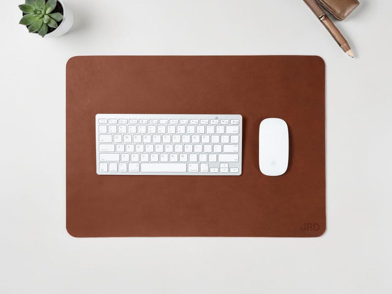 May include: A brown leather desk mat with a white keyboard and mouse. A small succulent plant in a white pot is in the upper left corner. The initials "JRD" are embossed in the lower right corner.
