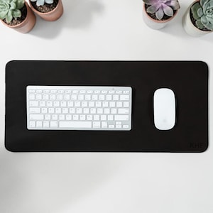 May include: A black rectangular desk mat with a white keyboard and mouse. The mat has rounded corners and the initials "KHF" in the bottom right corner. Small potted succulents are arranged around the desk mat.