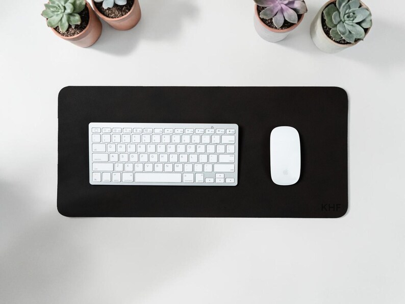 May include: A black rectangular desk mat with a white keyboard and mouse. The mat has rounded corners and the initials "KHF" in the bottom right corner. Small potted succulents are arranged around the desk mat.