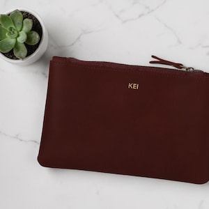 Pode incluir: Uma clutch de couro bord&ocirc; com fecho de z&iacute;per e as iniciais douradas "KEI". Uma pequena planta suculenta em um vaso branco &eacute; vis&iacute;vel. A clutch retangular est&aacute; sobre uma superf&iacute;cie de m&aacute;rmore branco.