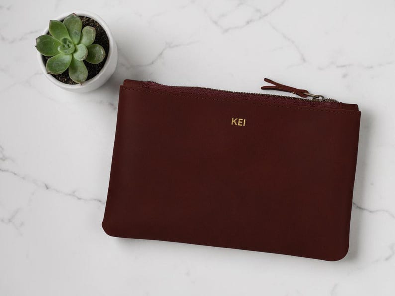 Pode incluir: Uma clutch de couro bord&ocirc; com fecho de z&iacute;per e as iniciais douradas "KEI". Uma pequena planta suculenta em um vaso branco &eacute; vis&iacute;vel. A clutch retangular est&aacute; sobre uma superf&iacute;cie de m&aacute;rmore branco.