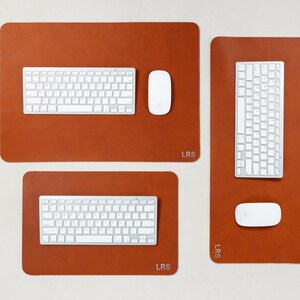 May include: Three brown leather desk pads with white keyboards and mice. The pads are rectangular with rounded corners. The top pad is the largest, with a keyboard and mouse. The other two pads have a keyboard and mouse. The pads have the initials "LRS".