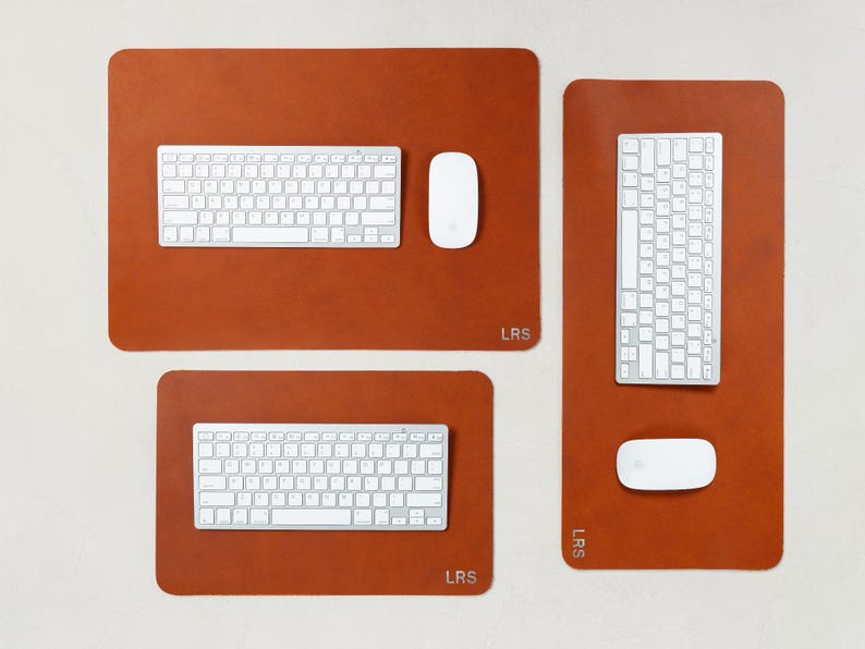 May include: Three brown leather desk pads with white keyboards and mice. The pads are rectangular with rounded corners. The top pad is the largest, with a keyboard and mouse. The other two pads have a keyboard and mouse. The pads have the initials "LRS".