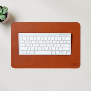 May include: A rectangular, brown leather desk mat with a white keyboard on top. The mat has rounded corners and the initials "LRS" in the bottom right corner. A small potted succulent is in the upper left corner.