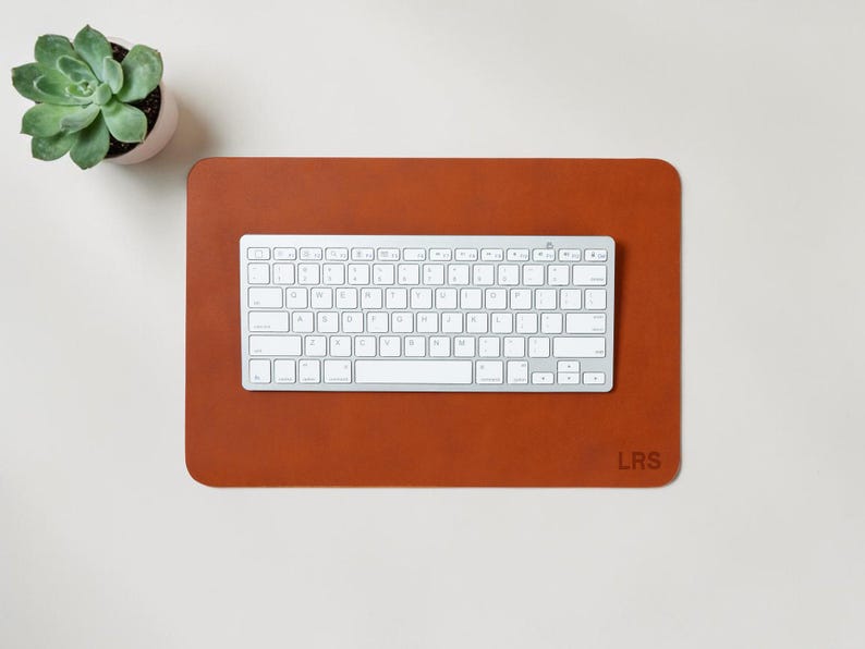 May include: A rectangular, brown leather desk mat with a white keyboard on top. The mat has rounded corners and the initials "LRS" in the bottom right corner. A small potted succulent is in the upper left corner.