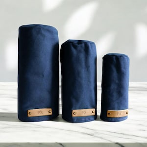 May include: Three navy blue golf club head covers of varying heights, each featuring a rectangular tan leather patch at the base. The leather patches are secured with two metal rivets and are embossed with the letters PX in a simple font.