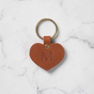Puede incluir: Un llavero de cuero marrón en forma de corazón con un llavero dorado. La letra "M" está grabada en el corazón. El llavero está sobre una superficie de mármol blanco.