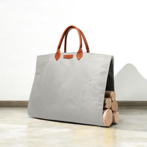 May include: A large, pale grey canvas tote bag with brown leather handles and details. The bag is filled with several cut logs, suggesting its use for carrying firewood. The bag is designed with a rectangular shape and a sturdy construction.