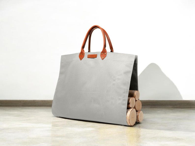 May include: A large, pale grey canvas tote bag with brown leather handles and details. The bag is filled with several cut logs, suggesting its use for carrying firewood. The bag is designed with a rectangular shape and a sturdy construction.